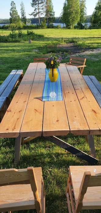 Utomhus träpicknickbord med bänkar och en gul kanna med vilda blommor på en löpare, med en lantlig utsikt.