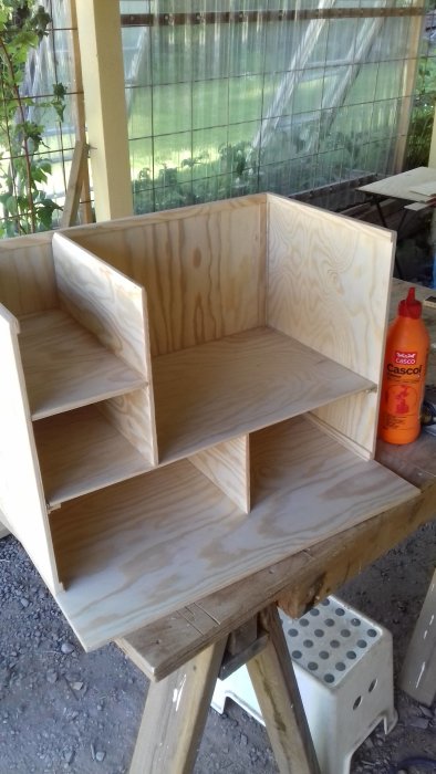 En halvfärdig träverktygslåda med hyllor, står på ett arbetsbord, limflaskan till höger.