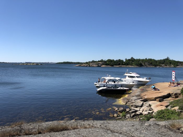 Två båtar förtöjda vid klippkant i skärgården, människor njuter av solen nära vattnet.