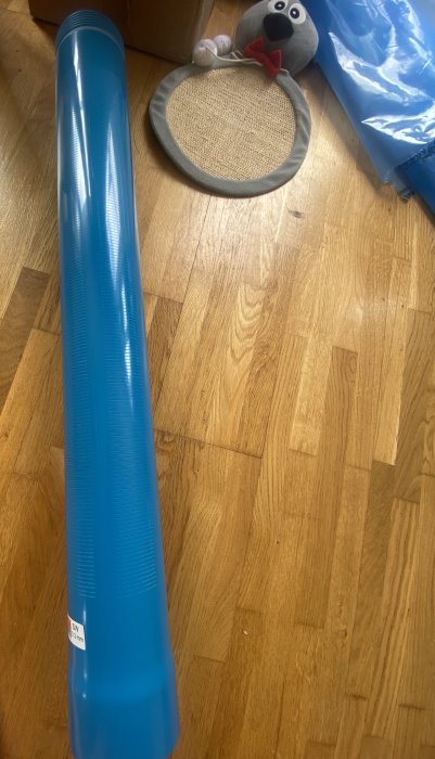 Blå plastmuff liggande på ett trägolv med en kattleksak i bakgrunden.