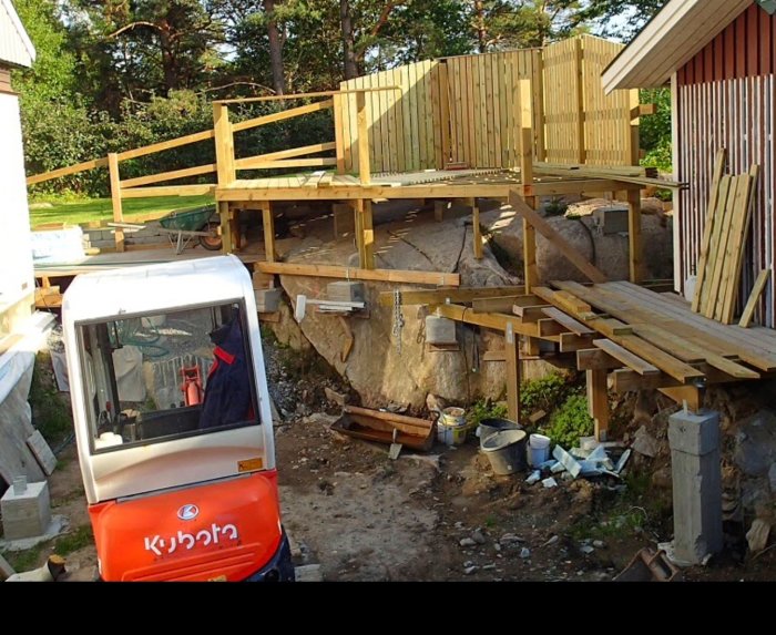 Byggplats med pågående konstruktion av trädäck och plintar, material och verktyg synliga, inklusive en minigrävare.