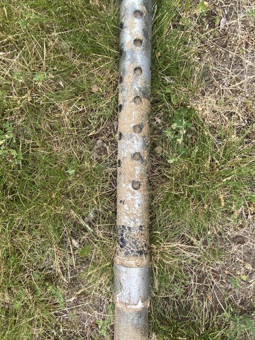 En bild av ett vertikalt borrat brunnsrör med flera hål, några igentäppta med sand, på en gräsmatta.