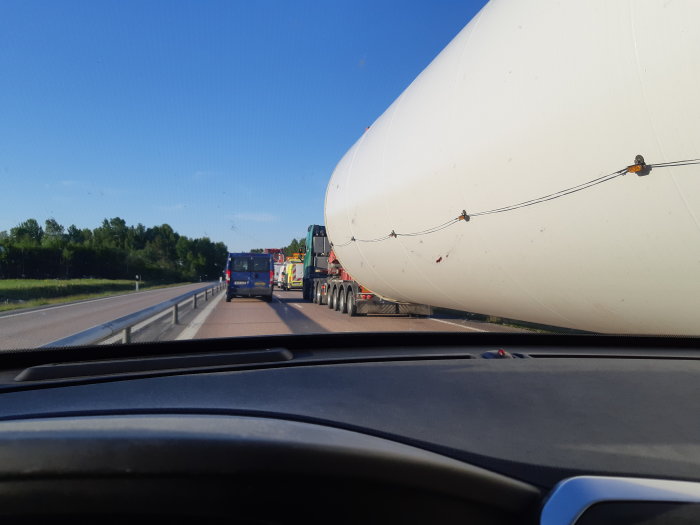 Lastbil som transporterar en stor vit cylinderformad last säkrad med spännband, sett från en annan bil.