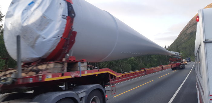 Vindkraftverksvinge transporteras på släpvagn längs väg med skog och berg i bakgrunden.