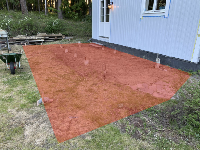 Markberedningsområde för trall intill en husfasad med avlägsnade plintar och verktyg i förgrunden.