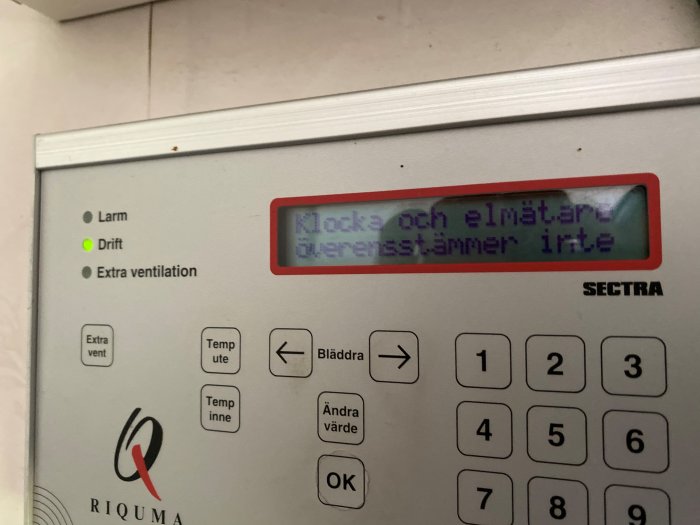 Kontrollpanel för ventilationssystem där larmindikatorn lyser och skärmen visar "Klocka och elmätare överensstämmer inte".