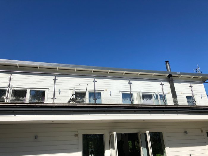 Vit modern villa med svart tak och regnränna, skorsten samt fönster med spegelglas under en klarblå himmel.