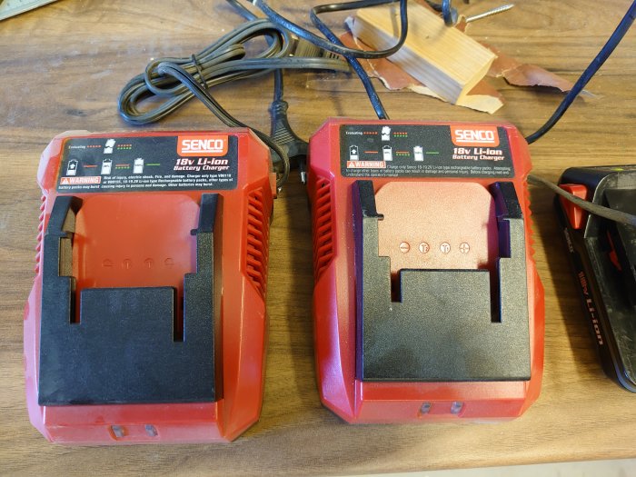 Två liknande röda SENCO 18V Li-Ion batteriladdare på ett träbord, den vänstra med elkabel ansluten.