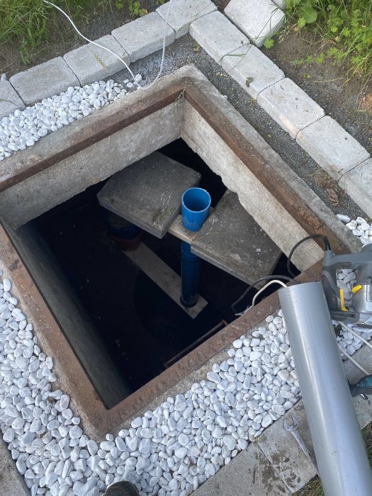 Öppen brunn med synbart rör som har sänkts ned, omgiven av vita stenar och betongblock.
