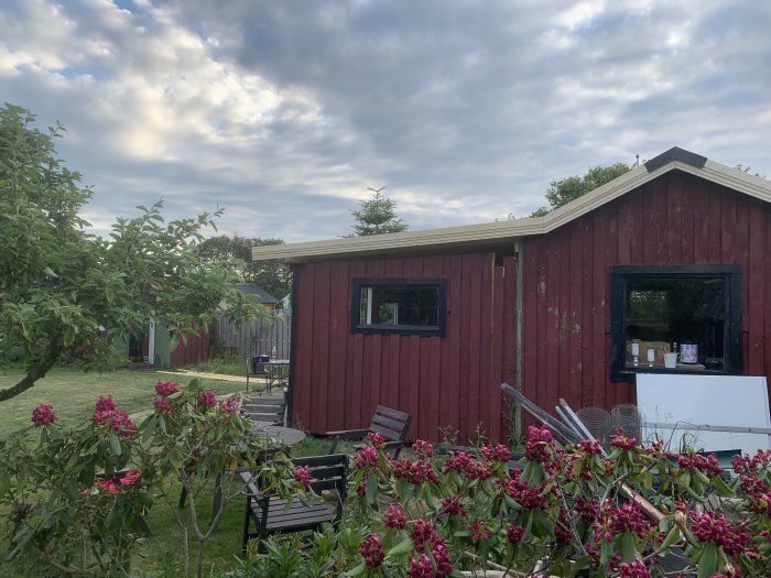 Ett rött trähus med nylagda vindskenor, omgivet av grönska och blommande växter i en trädgård.