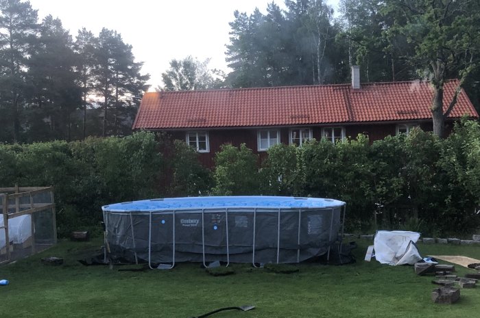 Ovanmark-pool i trädgård nära tomtgräns med röd stuga i bakgrunden.