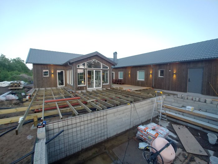 Pågående byggarbete med grunden för en tillbyggnad, inklusive utgrävd matjord, reglar, betongplattor och bärlinor.
