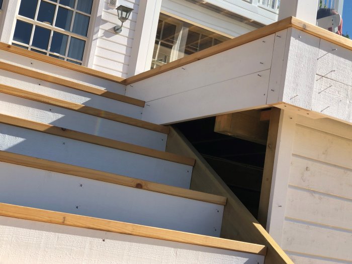 Del av en trätrappa med synlig bärlina under trappans överhäng, nära en husvägg.