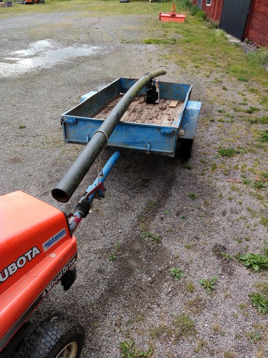 En kompakt traktor kopplad till en blå släpvagn med smuts och ett rör i släpvagnen.