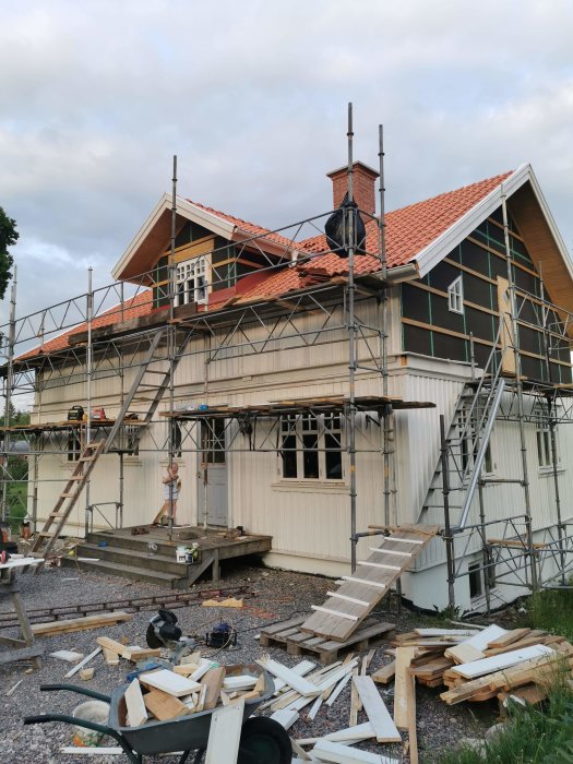 Hus under renovering med uppsatt panel på övervåningen, byggnadsställningar och byggmaterial på marken.