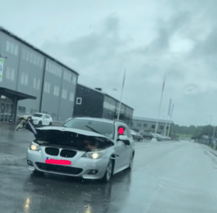 Bil med öppen bagagelucka där en persons ben sticker ut, på en våt väg utanför byggnader.