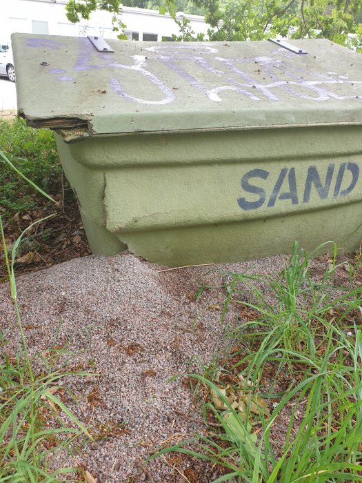 Grön sandlåda med sprucken kant där sand läckt ut på grusunderlag.