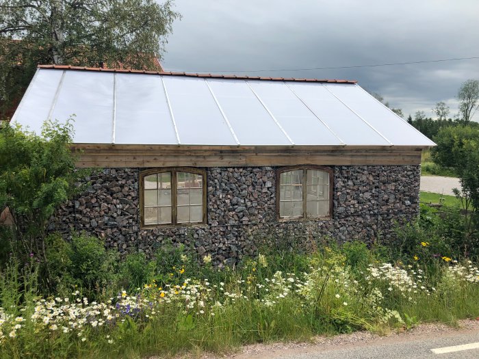 Växthus med gabionkorgar som stomme och oputsade stenväggar, vit takduk, omgivet av vilda blommor.