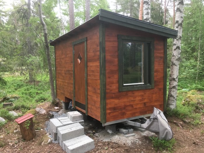 Liten stuga med träbeklädnad nästan färdigställd i skogsmiljö, väntar på ytterligare målning och vindskivor.