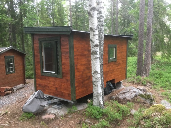 Nästan färdigställd timmerstuga med rödmålad panel och mörk taklist i skogsmiljö, skyddsplåten synlig vid hörn.