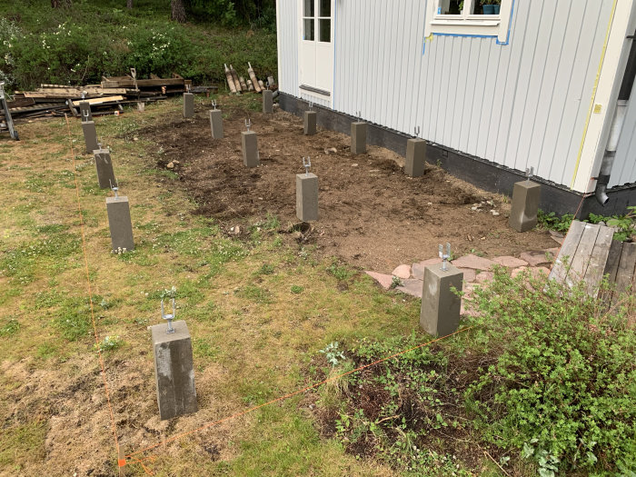 Utplacerade betongplintar för byggnation nära husvägg, markerat område för kommande trädäck.