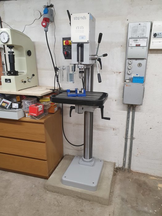 En pelarborrmaskin står stadigt på en betongplatta i ett verkstadsrum med elfördelare och verktyg i bakgrunden.