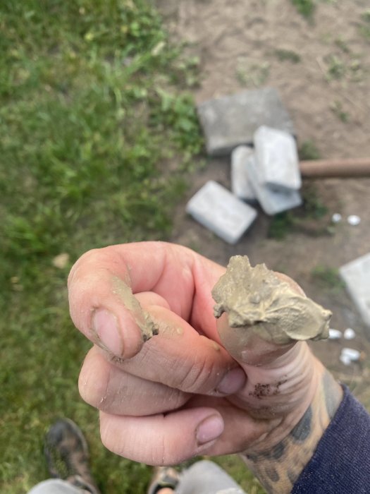 Hand visar jämförelse mellan fastklädd lera och sand med suddig bakgrund av gräsmark och grå betongplattor.