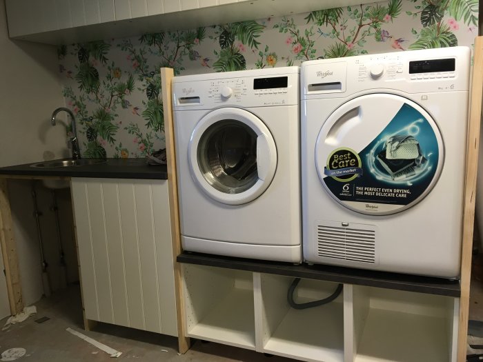 Tvättmaskin och torktumlare installerade under arbetsbänk med lös bänkskiva och tapet med blommönster.