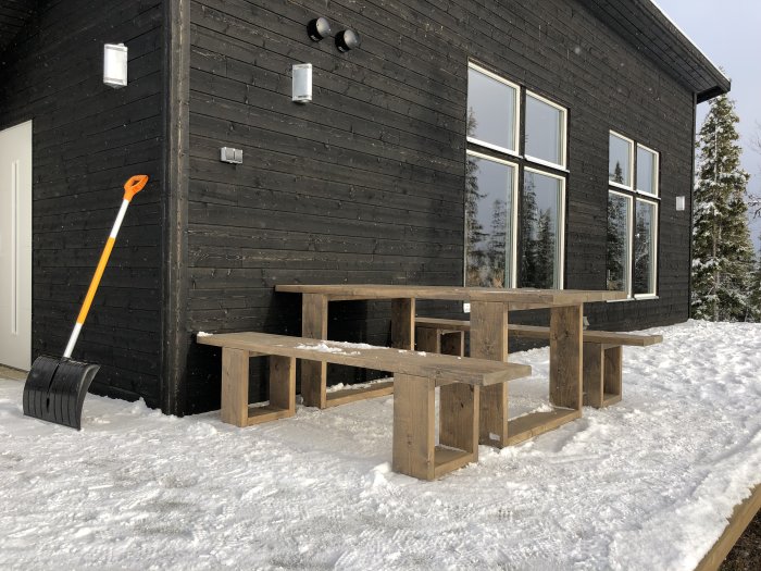 Svart trähus med stora fönster, en träsittgrupp ute i snön och en snöskyffel lutad mot väggen.