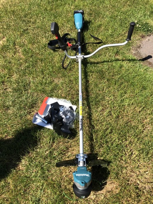 Batteridriven Makita DUR368AZ grästrimmer på gräs med kantklipparens tillbehör bredvid.