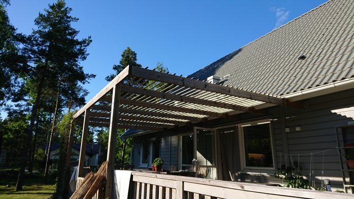 En nyinstallerad solskyddstrall monterad ovanpå pergola vid en husfasad, med trädgård och klarblå himmel i bakgrunden.