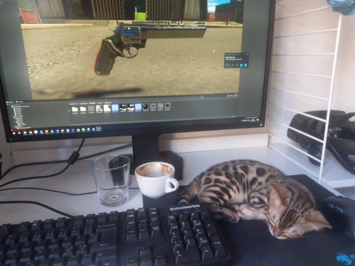 Kattunge somnar på skrivbord bredvid tangentbord, en tom kaffekopp och datorskärm visande ett spel.