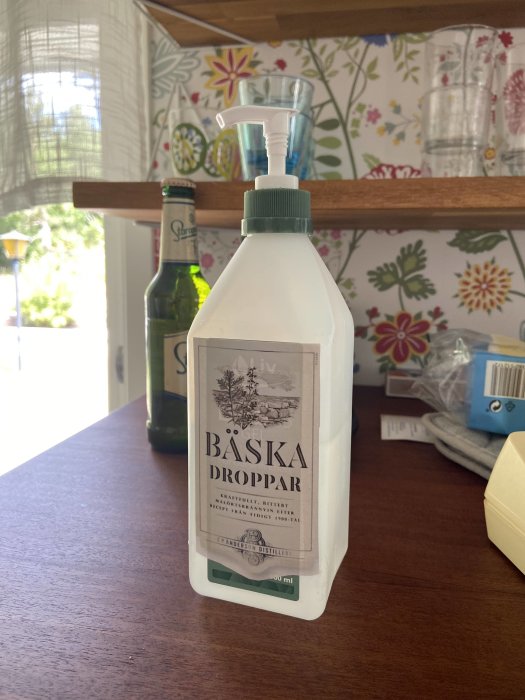 Flaska med Bäska Droppar framför en ölflaska och blommigt tyg i bakgrunden.