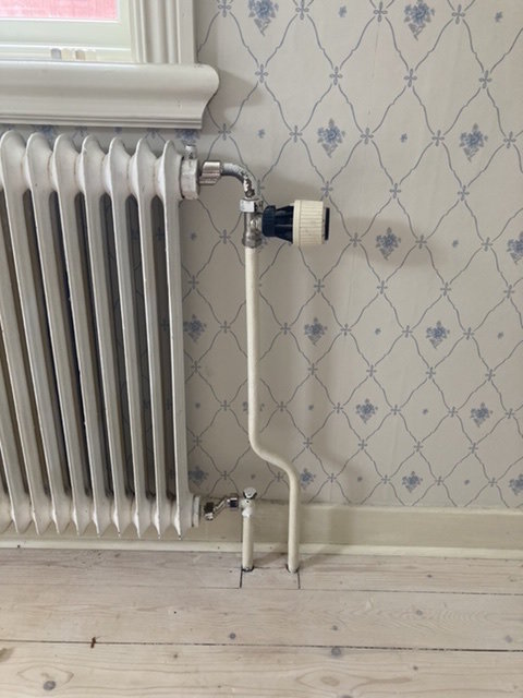 Vit radiator med justerad höjd tack vare två excenterkopplingar vid anslutning av rör, mot blommigt tapetserad vägg.