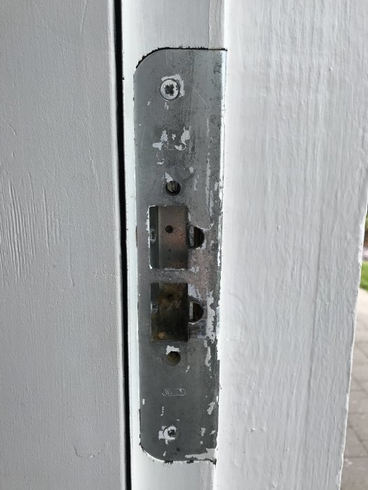 Närbild av ett dörrbleck med skruvar och slitage, monterat på vitmålad dörrkarm.