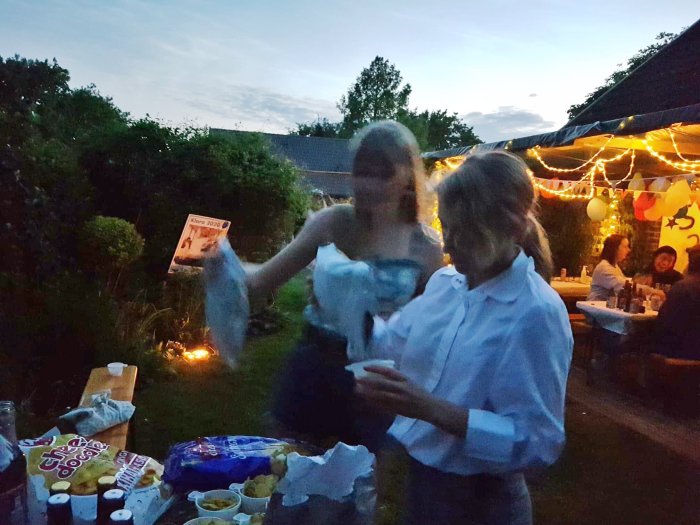 Kvällsparty i trädgården med suddiga personer, mat på bord och ljusslingor.