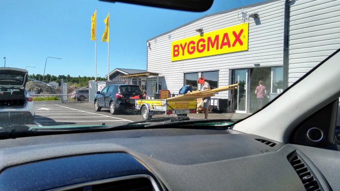 Bild på överlastat släp med träplank framför en Byggmax-butik en solig dag.