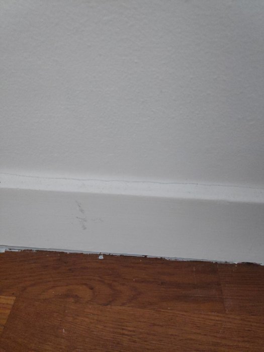 Skadad dörrlist och slitet golv med gammal parkett och smyglist vid väggkanten.