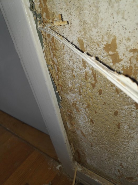 Skadat hörn av ett rum med flera lager bortskrapad tapet på vägg och gummitätning ovanpå list mot golvet.