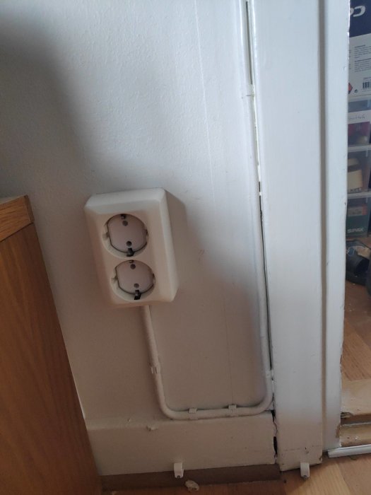 Ett gammalt dubbelt eluttag längs golvet med synliga yttre kablar och rör, nära en vit dörrpost.