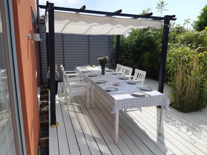 Nybyggd uteplats med matbord klädd i rutig duk, stolar och en pergola, redo för midsommarfirande.