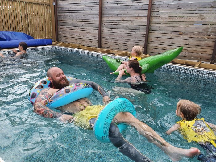 Personer firar midsommar med bad i en trädgårdspool, flytringar och leenden.