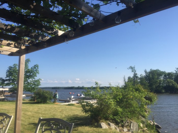 Trädgårdspergola med robusta stolpar och takbjälkar över en altan med utsikt över sjö och båtar.