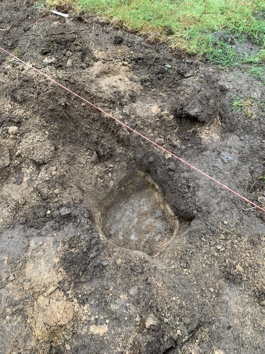 En upphöjd vy över en grävd grop som exponerar en sluttande bergsyta i en trädgård, markerad med ett rosa snöre.