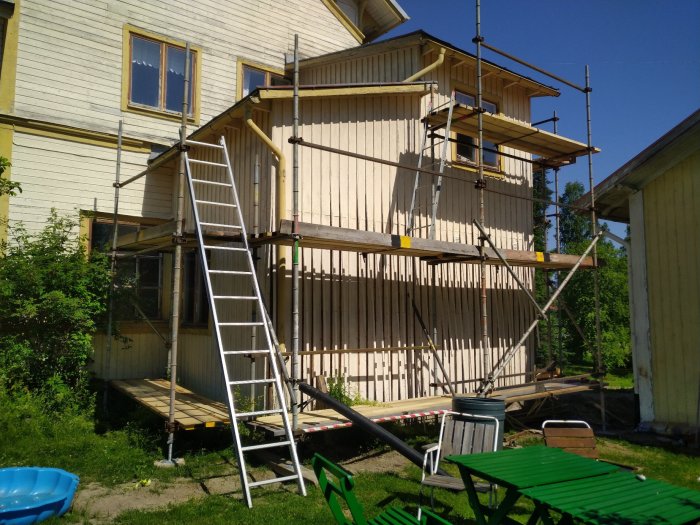 Byggställning framför ett hus under fasadrenovering med stege och trädgårdsmöbler i förgrunden på en solig dag.