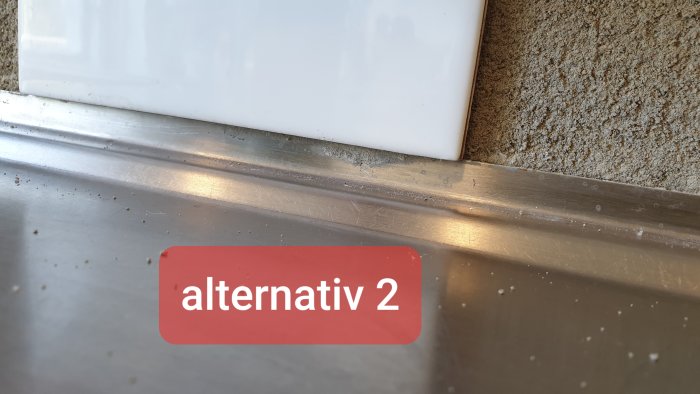 Närbild på en köksbänk med en metallist som möter en vit kakelplatta och textetiketten "alternativ 2".