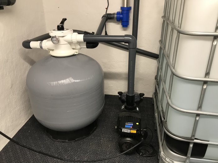 Teknikrum med filtertank, pump och IBC-vattentunnor på gummimatta mot vit vägg.