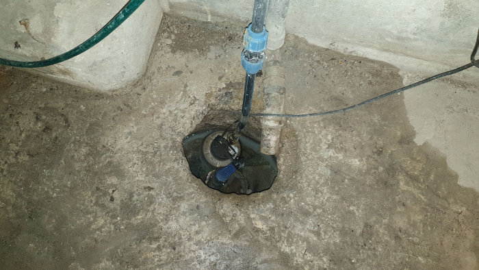 Dräneringspump i ett hål i en källare med en avkopplad avloppsrör som sticker ner och en slang som leder ut.