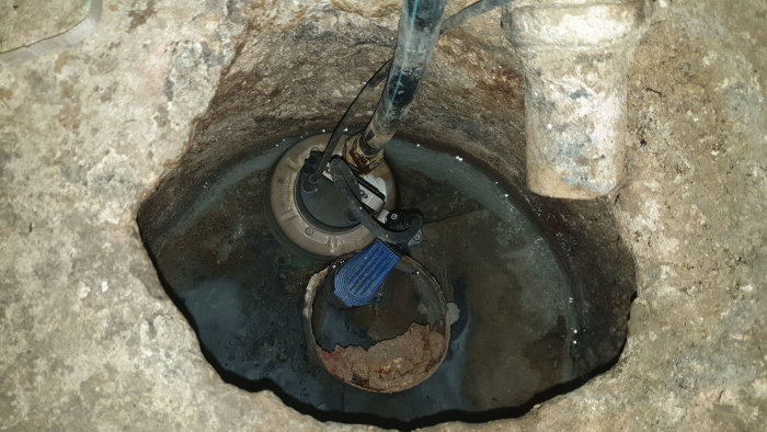 Ett hål i källaren med en dräneringspump och frigolitskiva, avloppsslang in i krypgrunden, oanslutet rör ovanför.