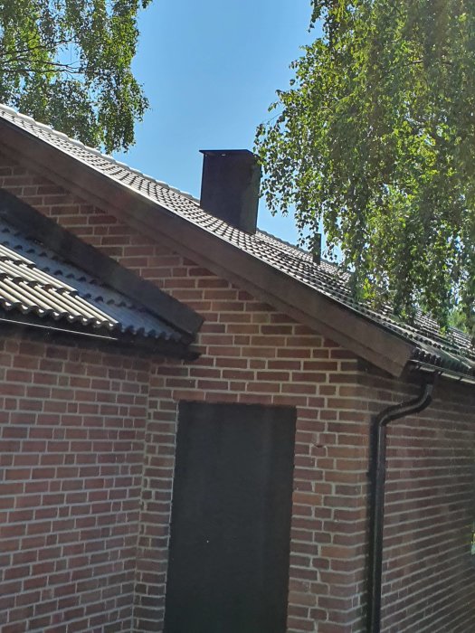 Bild på ett tegelhus med ett svart eternitventilationsrör på taket före byte till spiro, omgiven av gröna träd.